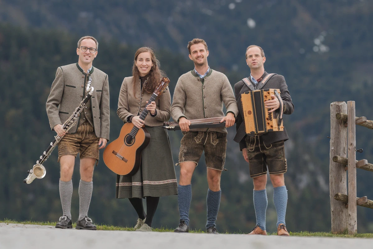 Die vier Musikant:innen der Almstüberl Musi stehen in Tracht nebeneinander vor einem Bergwald, mit Bassklarinette, Gitarre, Zither und Steirischer Harmonika